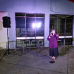 A woman stands outside the abandoned shell of a deserted library telling jokes at night